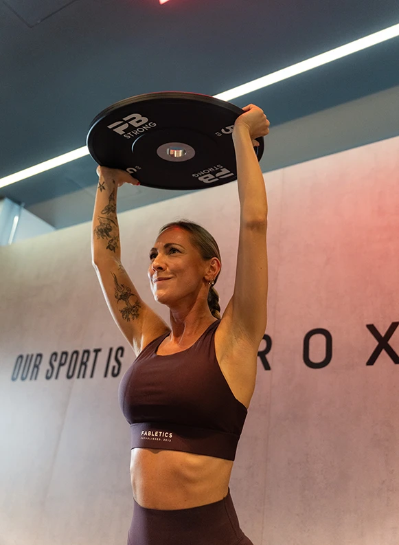 Frau stemmt Gewichtsscheibe über den Kopf im Trainingsbereich HYROX.