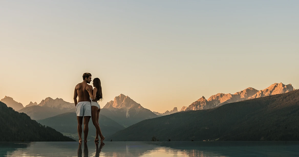 Mann und Frau in weißer Badekleidung stehen am Rande eines Hotelpools, im Hintergrund die massive Bergwelt der Dolomiten im Sonnenaufgang.