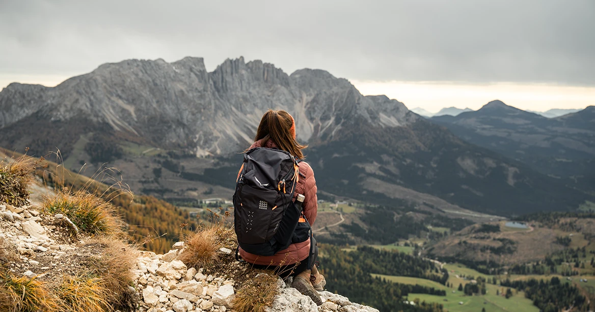 Frau mit Rucksack sitzt auf einem Stein auf einem Berg und schaut auf ins davor liegende Tal, dahinter ragt ein großes Felsmassiv der Dolomiten empor.