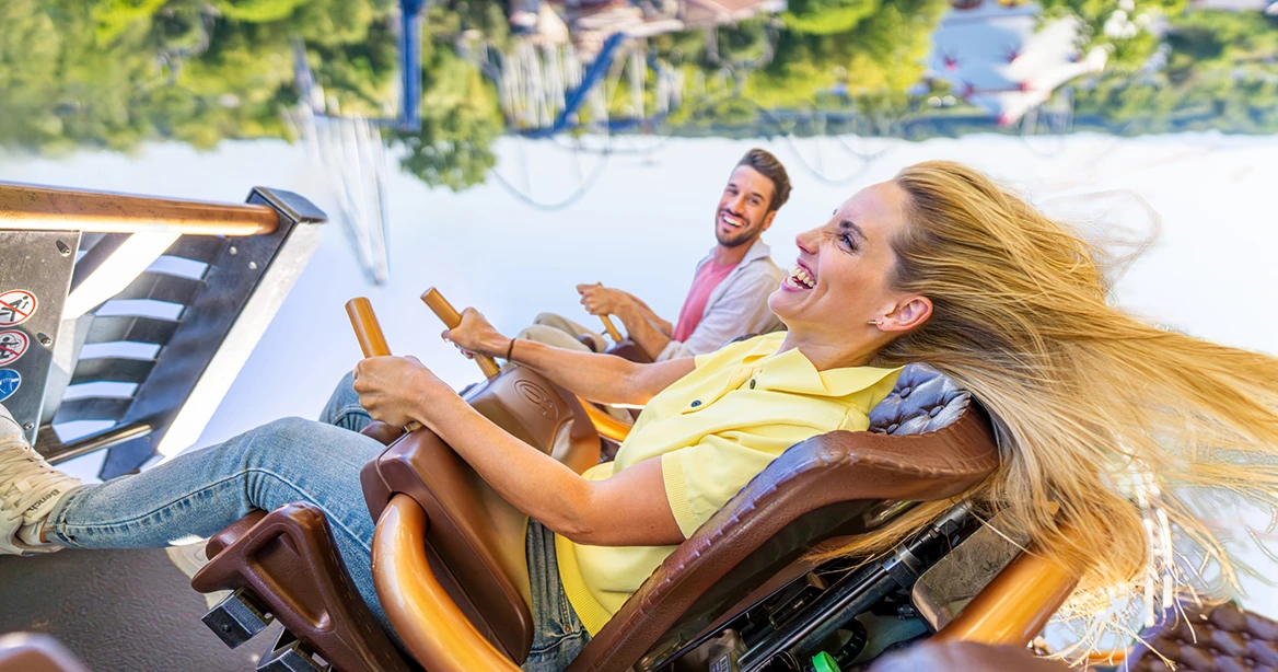 Eine lachende junge Frau mit fliegenden Haaren und ihr Partner sitzen zusammen in einer Achterbahn.