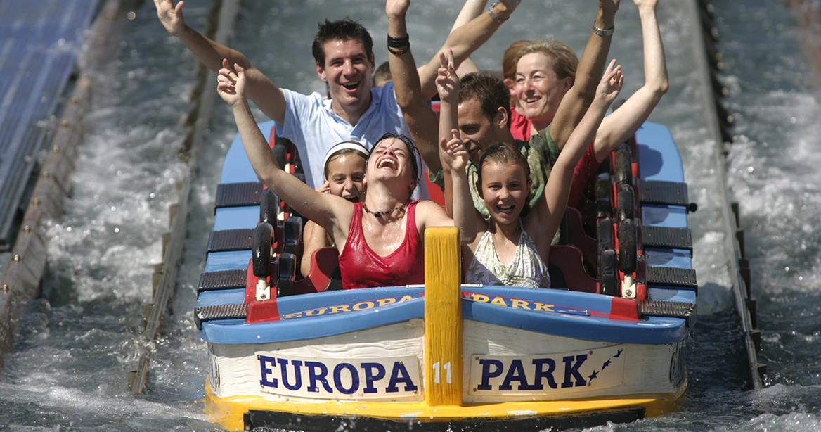 Im Europa-Park saust eine Gruppe nasser Menschen in einem Boot die Poseidon-Wasserachterbahn hinab. Alle reißen die Arme hoch.
