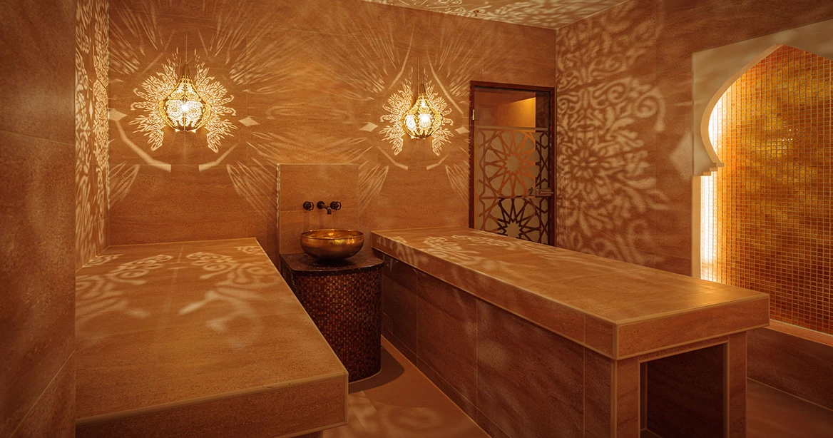 Zwischen 2 gefliesten Hamam-Liegen befindet sich an der Wand ein Waschbecken im Oriental Look. An der Wand hängen über dem Kopfende der Liegen zwei marokkanische Messingleuchten, die für Lichtspiele an der hellbeige gekachelten Wand und auf den Liegen sorgen.