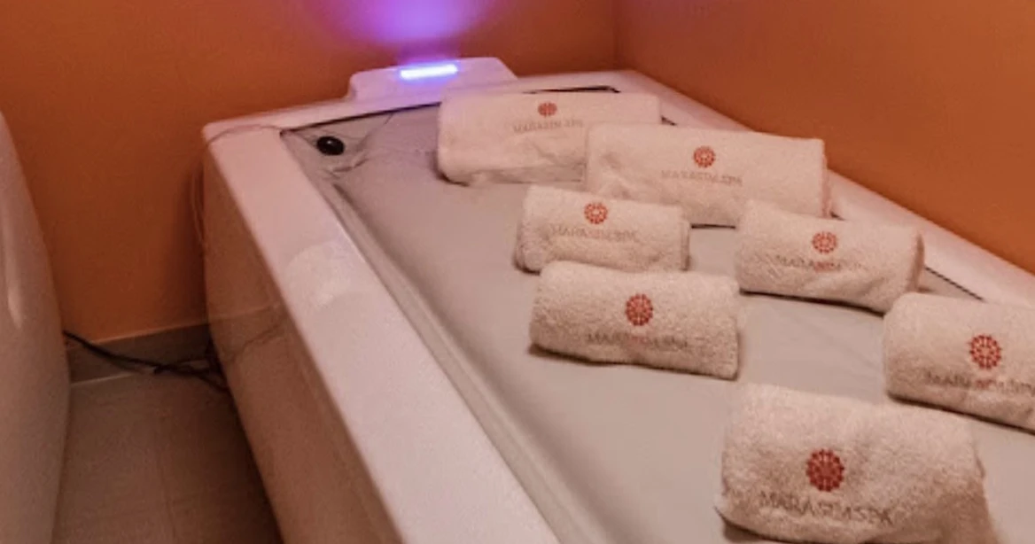 Auf einer elektrisch verstellbaren Massageliege liegen aufgerollte und ordentlich aufgereihte weiße Handtücher mit Marasim Spa Logo.