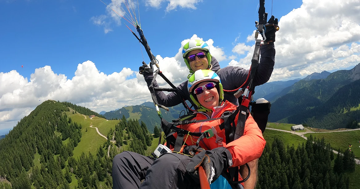 Nahaufnahme Mann und Frau im Gleitschirm-Tandemflug, im Hintergrund die bayerische Berglandschaft mit weißen Wattewolken an einem strahlend blauen Himmel.