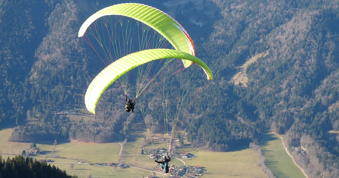 2 Gleitschirmflieger mit 2 giftgrünen Gleitschirmen segeln über ein sonnenbeschienenes Tal im bayerischen Voralpenland.