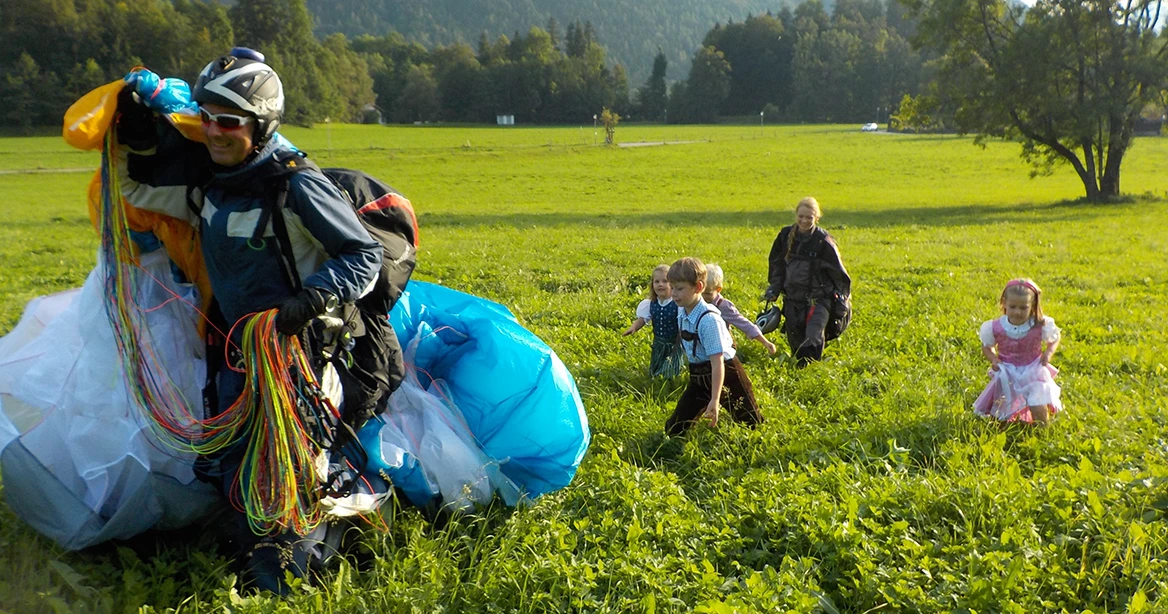 Ein gerade eben gelandeter Fallschirm-Flieger geht mit seinem zusammengerafften Fallschirm über dem Rücken, und gefolgt von einer Schar Kinder in Lederhosen und Dirndl sowie einer weiteren Fallschirm-Fliegerin, durch das hohe Gras auf einer Wiedse.
