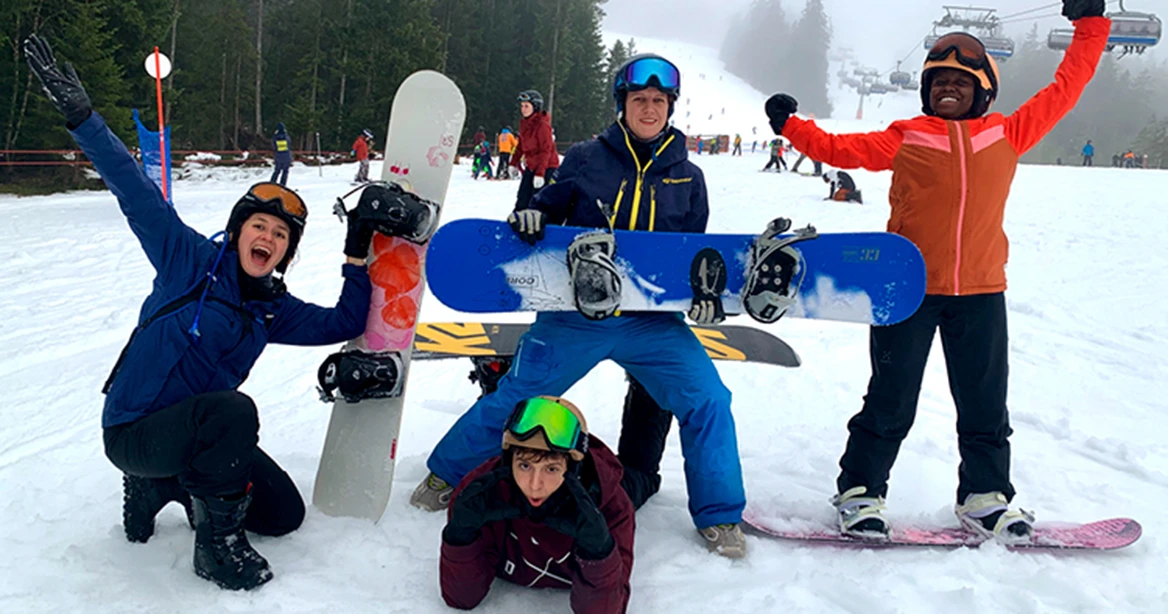4 lachende Snowboarder in bunten Skijacken und in einer artistischen Aufstellung am unteren Ende einer Skipiste.