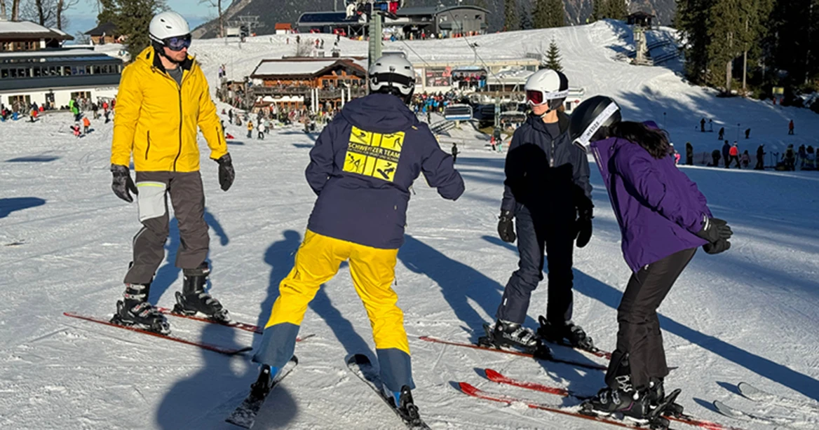 Ein Skilehrer und drei Schüler auf Skiern, im Hintergrund Skihütten und Skilift.