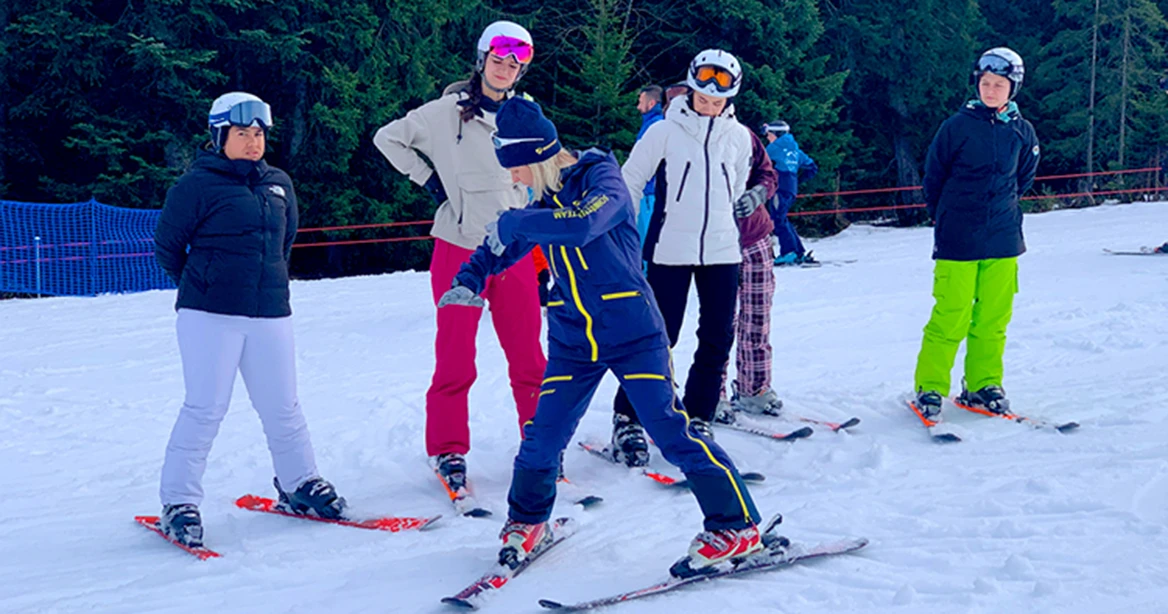 Eine Skilehrerin demonstriert den Schneepflug und die Drehung vor ihren Schülern auf einem Skihang.