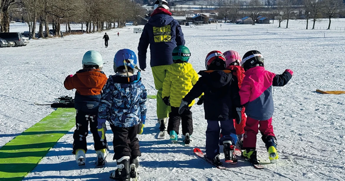 Ski-Kindergarten: 6 kleine Skifahrer folgen ihrem Skilehrer. Jeder trägt nur einen Ski am Fuß.