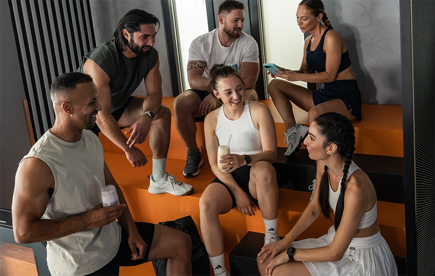 Drei Männer und drei Frauen sitzen nach dem Workout entspannt zusammen im Fitnessstudio. Einige haben Shakes in der Hand, während eine Frau den anderen etwas auf ihrem Handy zeigt.