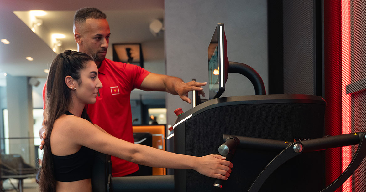 Trainer im Teamoutfit erklärt Übung am EGYM Gerät