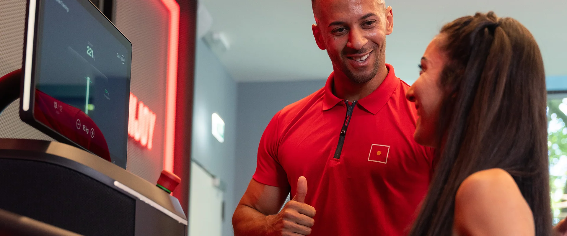 Eine Frau trainiert am EGYM-Gerät, während ihr Trainer sie mit einem motivierenden Daumen hoch unterstützt.