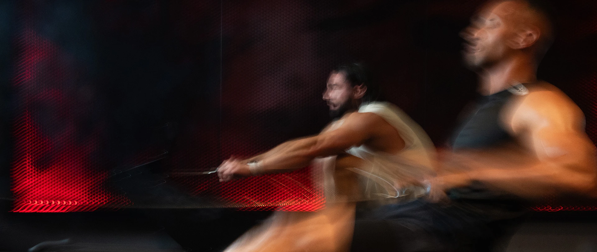 Zwei Männer trainieren auf dem Rower. Das Bild fängt die Bewegung ein und ist dadurch leicht blurry.