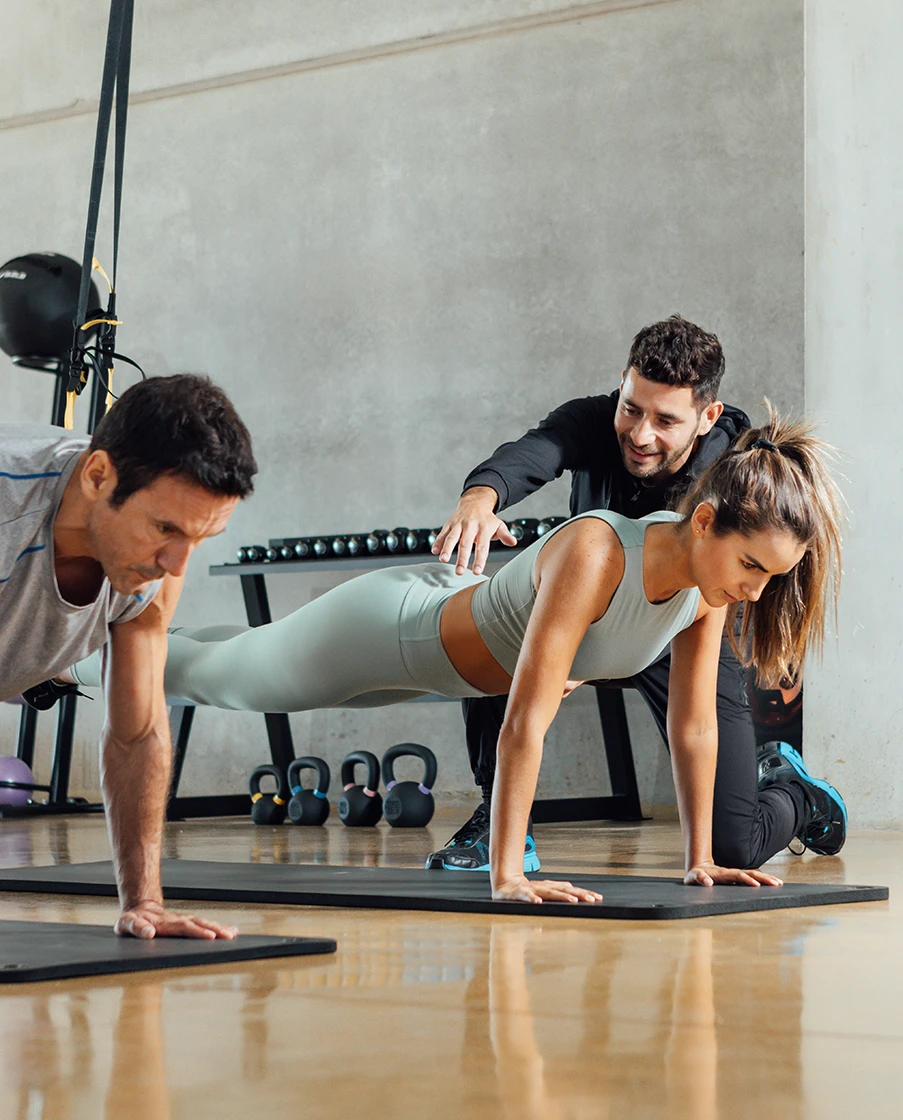 Ein Trainer unterstützt eine Frau bei der Ausführung einer Plank-Übung im Fitnessstudio. Zusätzlich zeigt das Bild einen trainierenden Mann bei der gleichen Übung in einer Detailaufnahme.