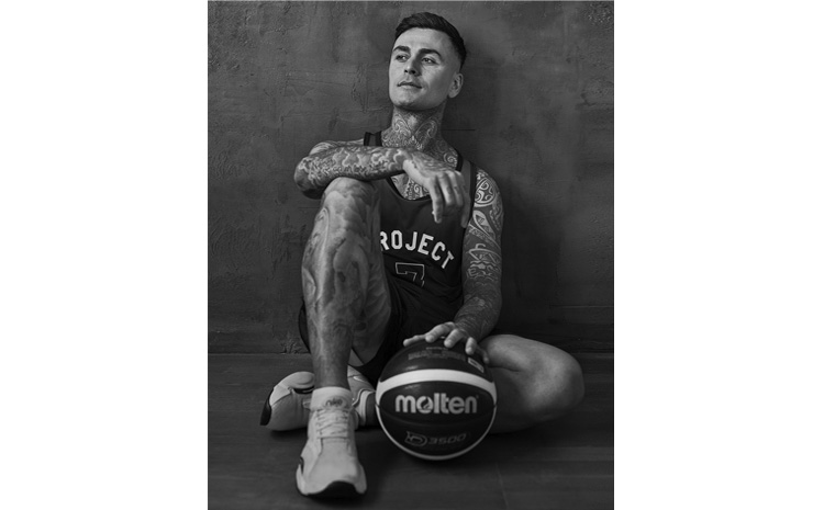 Junger Mann im Basketball-Outfit sitzt auf dem Boden mit dem Rücken an der Wand, vor ihm ein Basketball.